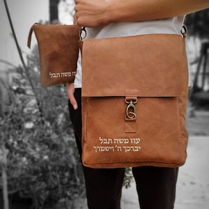 May include: A brown leather messenger bag with a silver clasp and a smaller brown leather pouch attached. The bag has white embroidered text in Hebrew that reads "עוד מעט הכל יהיה בסדר" which translates to "Soon everything will be alright".