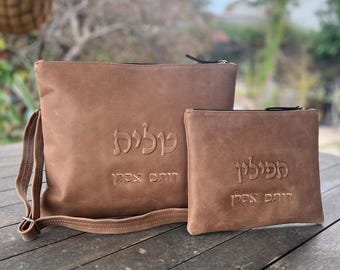 Puede incluir: Dos bolsas de cuero marrón con cremalleras. La bolsa más grande tiene una correa para el hombro y el texto "קוליה" y "דום אבינו" en relieve en la parte delantera. La bolsa más pequeña tiene el texto "מקוליה" y "דום אבינו" en relieve en la parte delantera.