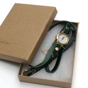 May include: A gold-tone watch with a white face and black hands is attached to a green leather strap. The watch is in a brown cardboard box with the word "Sigal Levi" printed on the top.
