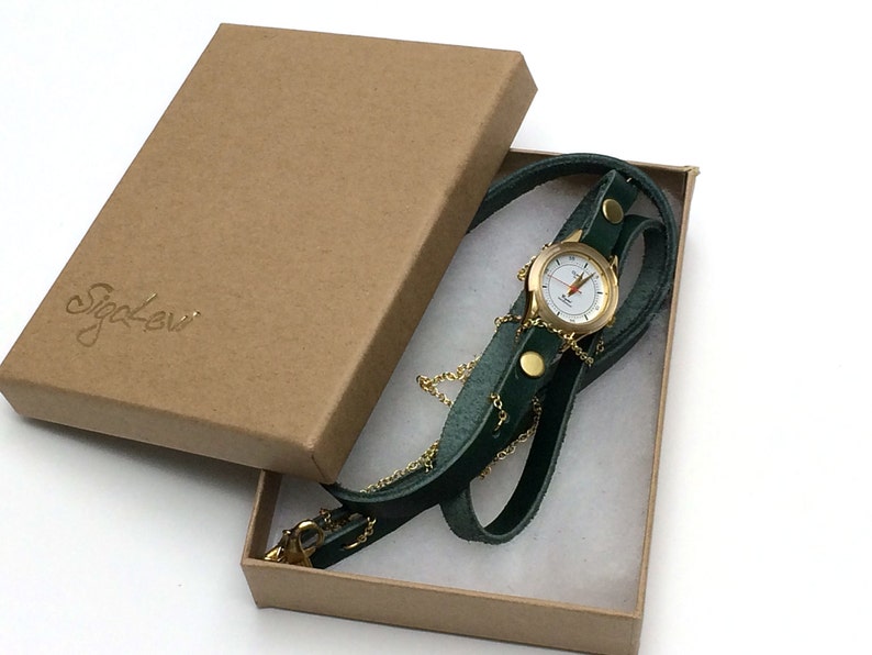May include: A gold-tone watch with a white face and black hands is attached to a green leather strap. The watch is in a brown cardboard box with the word "Sigal Levi" printed on the top.