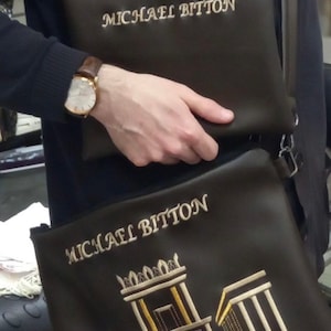 May include: Two brown leather bags with gold embroidery. The top bag has the name "MICHAEL BITTON" embroidered on it. The bottom bag has the same name and a stylized image of a building with Hebrew text.