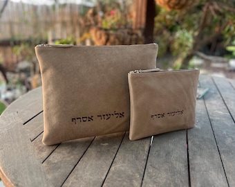 Puede incluir: Dos bolsas de gamuza color beige con cierres de cremallera se muestran sobre una mesa de madera. La bolsa más grande es rectangular y la más pequeña es cuadrada. Ambas bolsas tienen texto en color marrón oscuro.