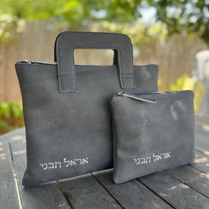 May include: Two gray leather bags with zipper closures and handles. The larger bag has a top handle and the smaller bag has a zipper pull. Both bags have Hebrew text embroidered on them.