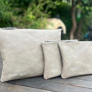 May include: A set of three light gray suede-like pouches with silver zippers. The largest pouch has text embroidered in a script font. The two smaller pouches also have embroidered text. The pouches are displayed on a wooden surface.