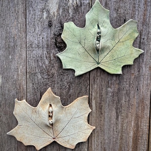 Puede incluir: Dos esculturas de cerámica con forma de hoja de arce con una cara tallada en el centro. Una hoja es verde claro, la otra beige. Ambas tienen patrones de venas detallados y están montadas sobre una superficie de madera envejecida.
