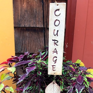 May include: A white ceramic sign with the word "COURAGE" written in brown lettering hangs from a black metal hook. The sign is attached to a leaf-shaped ceramic ornament. The sign is hanging in front of a wooden wall.