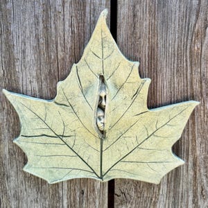 May include: A pale green, leaf-shaped ceramic wall plaque featuring a central vertical slit. Within this opening, two small, sculpted human faces are visible, one positioned above the other, with detailed facial features and a weathered texture.
