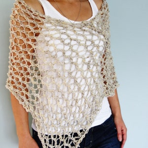May include: A beige crochet poncho with a loose, open weave. The poncho is worn over a white tank top and blue jeans.