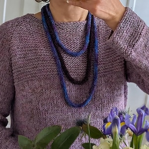 May include: A person wearing a lavender knit sweater and holding a bouquet of flowers. The bouquet includes white roses, purple irises, and green foliage. Two beaded necklaces in shades of blue and black are worn.