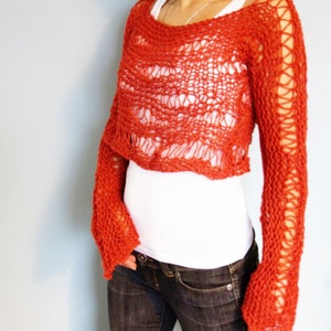 May include: A red crocheted sweater with a loose, open knit pattern. The sweater has long sleeves and a cropped length. The sweater is worn over a white shirt and blue jeans.