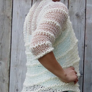 May include: A white crocheted cardigan with a loose, open weave. The cardigan has a slightly cropped length and three-quarter sleeves. The model is wearing the cardigan over a light blue shirt and blue jeans.