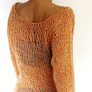 May include: An orange, open-weave knit top with long sleeves. The top has a scoop neckline and a loose, relaxed fit. The knit pattern creates a see-through effect, and the sleeves have a fringed detail at the cuffs. The top is worn with blue pants.