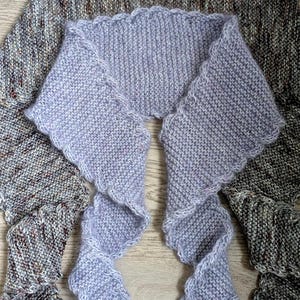 Könnte beinhalten: Ein zweiteiliges Strick-Set bestehend aus einem breiten, braun-grau gesprenkelten Schal und einem kleineren, einfarbigen helllavendelfarbenen Schal mit gewelltem Rand. Beide Teile weisen eine strukturierte Strickoptik auf.