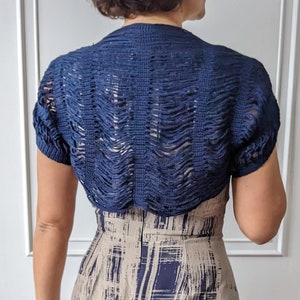Puede incluir: Un bolero de crochet azul marino con mangas cortas. El bolero está hecho de una tela delicada de tejido abierto y tiene un borde ligeramente festoneado.