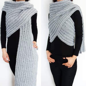May include: A gray knitted scarf with a long, flowing design. The scarf is worn in a wrap style, with one end draped over the shoulder and the other end hanging down the front of the body.