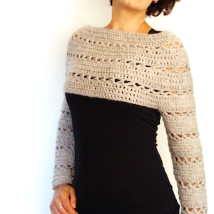 May include: A beige crochet shrug with a lacy pattern. The shrug has long sleeves and a wide neckline. The shrug is worn over a black long-sleeved shirt.