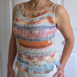 May include: A sleeveless, multi-colored crochet top with horizontal stripes in shades of orange, pink, blue, and yellow. The top has a scoop neckline and a slightly ruffled hem. The person is wearing white pants.