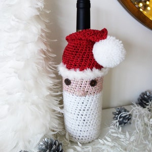 Puede incluir: Una botella de vino vestida de Papá Noel con un gorro rojo de ganchillo, un pompón blanco y una barba blanca de ganchillo. La botella está sentada sobre una superficie blanca con ramas de plumas blancas y piñas plateadas.