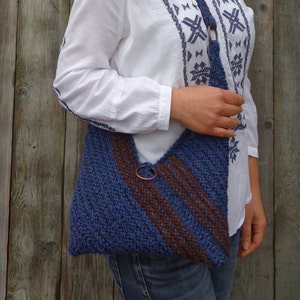 Może przedstawiać: Niebiesko-brązowa, dzianinowa torba na ramię z metalowym kółkiem. Torba jest noszona na ramieniu osoby w białej bluzie z niebiesko-białym wzorem śnieżynek.