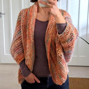 May include: A handmade, open-front crochet cardigan in shades of orange, peach, and blue. The cardigan has wide sleeves and a relaxed fit. The garment is made with a textured stitch, creating a cozy and stylish look. The cardigan is a perfect addition to any wardrobe.