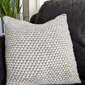 Op de afbeelding: Een vierkant, lichtgrijs gebreid kussen met een textuurpatroon. Het kussen ligt op een zwarte leren bank, met een groene plant zichtbaar op de achtergrond. Het ontwerp van het kussen heeft een herhalend patroon van verhoogde steken.