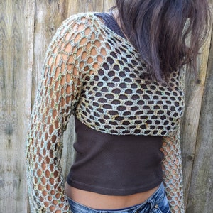 May include: A crocheted top with long sleeves in a green and brown color scheme. The top is a loose fit and has a mesh design. The model is wearing the top over a brown tank top and blue jeans.