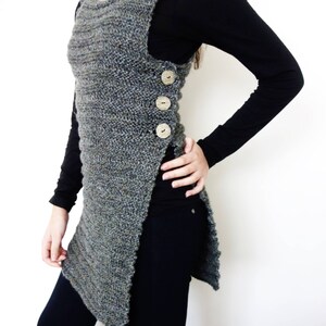 May include: A long, gray knitted vest with a side slit and buttons. The vest is worn over a black long-sleeved shirt and black pants.
