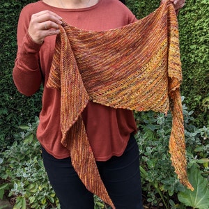 Puede incluir: Una chal de punto colorido con un degradado de naranja, amarillo y marrón. La chal está drapeada sobre los hombros de una persona y tiene un extremo largo y fluido.