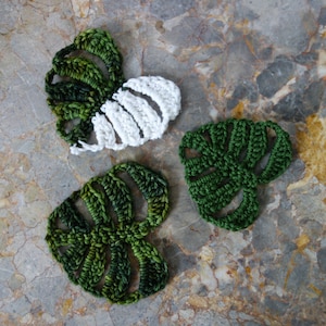May include: Three crocheted monstera leaves in shades of green and white. The leaves are made with a delicate stitch and have a realistic appearance.