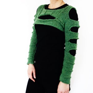 May include: Green knitted arm warmers with cutouts. The arm warmers are worn over a black long-sleeved shirt.