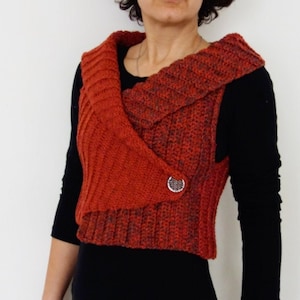 Può includere: Un gilet di colore ruggine lavorato a uncinetto con un ampio colletto e una chiusura con un solo bottone. Il gilet è corto e presenta un design leggermente asimmetrico.