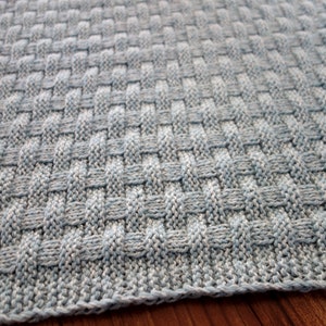 May include: A close-up of a knitted baby blanket in a light blue color with a woven pattern.