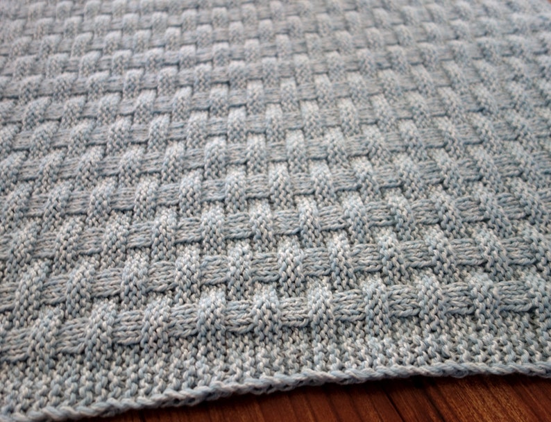 May include: A close-up of a knitted baby blanket in a light blue color with a woven pattern.