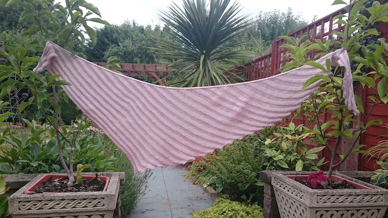May include: A pink knitted shawl with a striped pattern, draped over a bush in a garden setting.