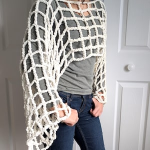 May include: A white crochet poncho with a large open weave pattern. The poncho is worn over a gray t-shirt and blue jeans.