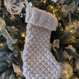 Pode incluir: Uma meia de Natal de malha branca pendurada em uma árvore de Natal decorada. A meia tem uma textura grossa e felpuda e uma bainha larga.