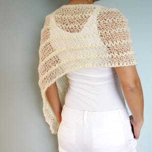 May include: A white crocheted shawl with a delicate lace pattern. The shawl is draped over the shoulders of a person wearing a white top and white pants.