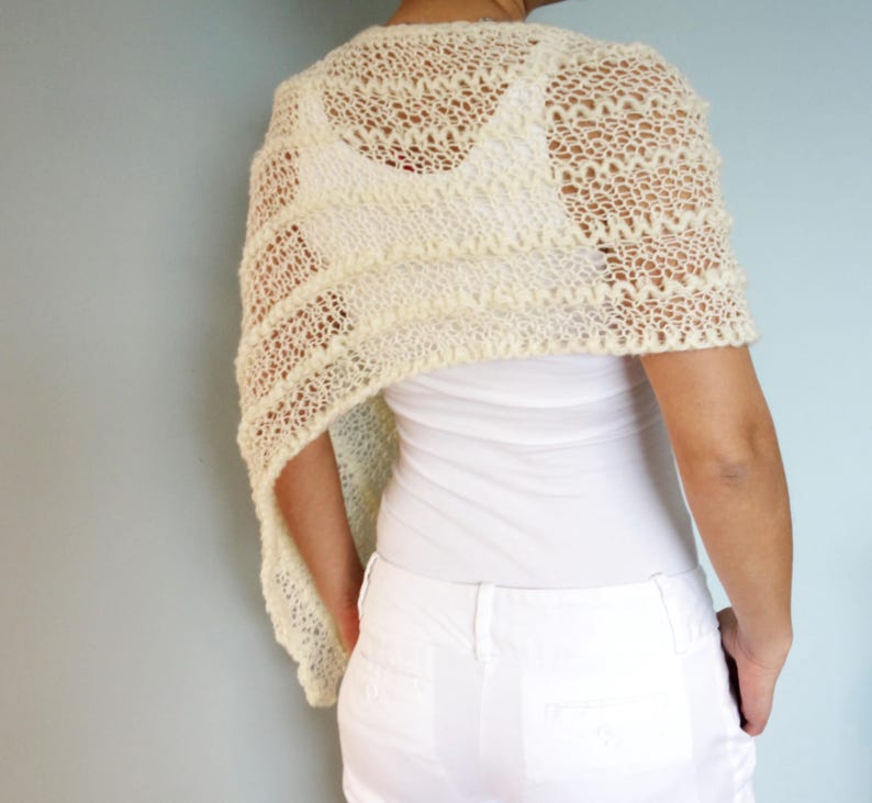 May include: A white crocheted shawl with a delicate lace pattern. The shawl is draped over the shoulders of a person wearing a white top and white pants.