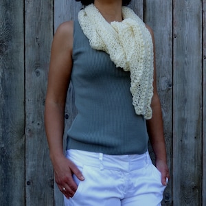 May include: A woman wearing a white crocheted scarf over a teal sleeveless top and white pants.