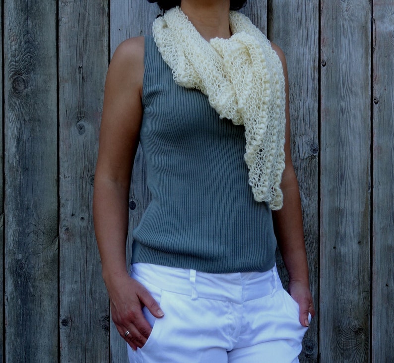 May include: A woman wearing a white crocheted scarf over a teal sleeveless top and white pants.