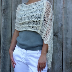 May include: A white crocheted shawl with a lacy pattern. The shawl is draped over a person's shoulders and is worn with a gray shirt and white pants.