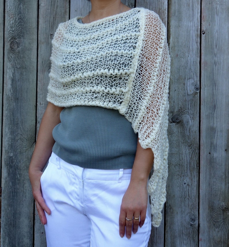 May include: A white crocheted shawl with a lacy pattern. The shawl is draped over a person's shoulders and is worn with a gray shirt and white pants.