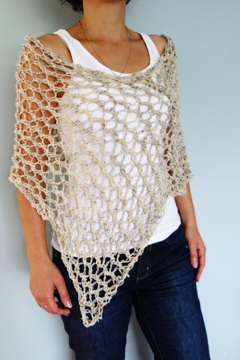 May include: A beige crochet poncho with a loose, open weave. The poncho is worn over a white tank top and blue jeans.