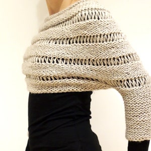 May include: A beige knitted shrug with a loose, open weave and a single long sleeve. The shrug is draped over the shoulders and has a relaxed fit.
