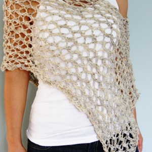 May include: A beige crochet poncho with a loose, open weave. The poncho is worn over a white tank top and blue jeans.