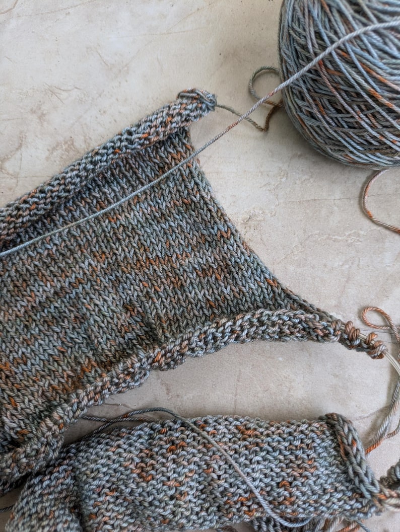 May include: Close-up of a knitting project in progress, featuring a section of knitted fabric in shades of gray, blue, and orange. A ball of yarn in matching colors sits nearby. The texture of the knitted stitches is clearly visible.