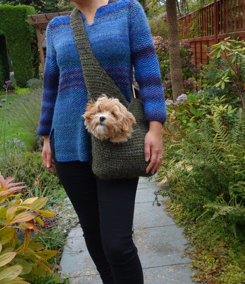May include: A woman wearing a blue and green striped sweater carries a small dog in a green knitted sling bag.