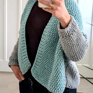 Puede incluir: Cárdigan tejido a mano en dos tonos. El diseño abierto presenta un cuerpo azul claro y mangas grises. La construcción de punto grueso es visible. El cárdigan se usa sobre una camiseta oscura y vaqueros azules.