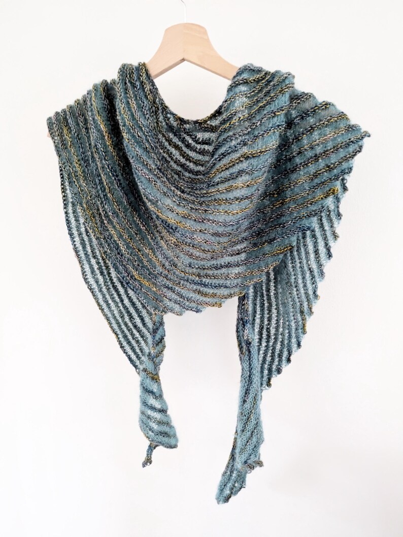 May include: A teal and gold striped knitted shawl with a fringed edge. The shawl is draped over a wooden hanger.