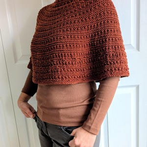 Pode incluir: Um poncho tricotado &agrave; m&atilde;o na cor ferrugem com um padr&atilde;o de malha canelada texturizada. A pe&ccedil;a tem um decote alto e arredondado e uma silhueta curta tipo capa que cai sobre os ombros, usada sobre uma camisa castanha de manga comprida.